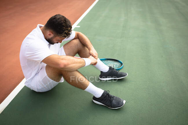 Ein Mann mit gemischter Rasse in weißen Tennisanzügen, der an einem sonnigen Tag auf einem Tennisplatz spielt, eine Pause macht und auf einem Boden sitzt — Stockfoto