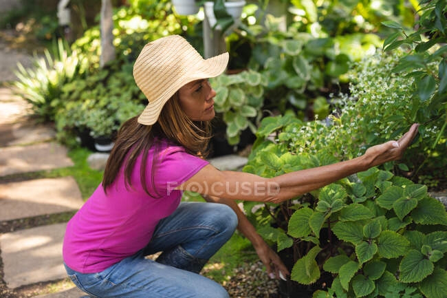 Una donna caucasica indossa una t-shirt rosa e un cappello di paglia, godendo del tempo in un giardino soleggiato, toccando le foglie delle piante. — Foto stock