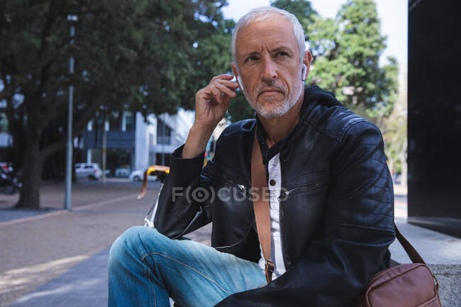 Älterer Mann aus dem Kaukasus, lässig gekleidet, tagsüber auf den Straßen der Stadt unterwegs, auf Treppen sitzend und mit Kopfhörern. — Stockfoto