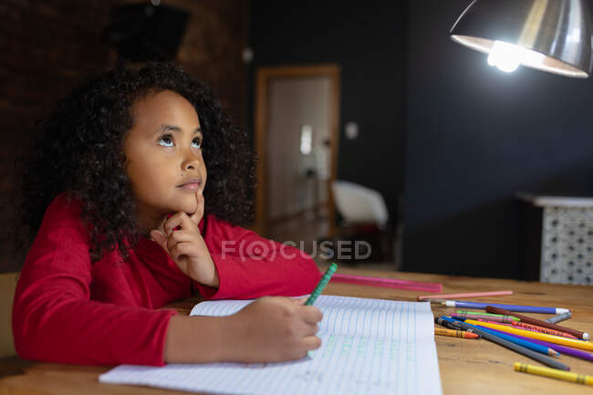 Вид сбоку на молодую афроамериканскую девушку дома, сидящую за обеденным столом, делающую домашнее задание, глядящую вверх в мыслях с открытой школьной книгой перед ней — стоковое фото