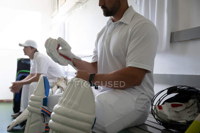 Side view mid section of a mixed race male cricket player wearing whites, sitting on a bench in a changing room, preparing to the game, putting on cricket gloves, with another player sitting in behind. — Stock Photo