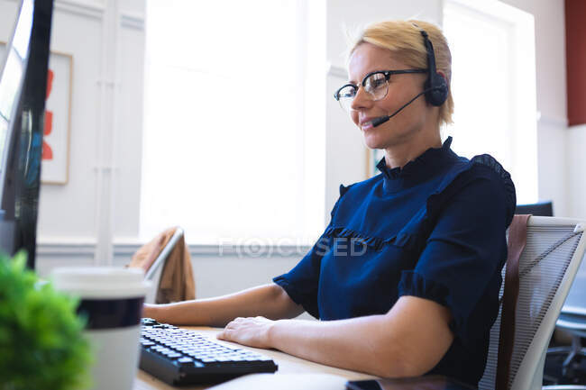 Una mujer de negocios caucásica con el pelo corto y rubio, trabajando en una oficina moderna, sentada en un escritorio, con auriculares y hablando - foto de stock