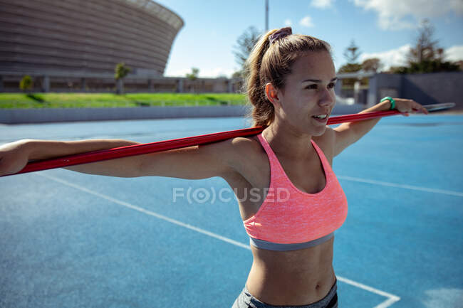 Side view of a relaxed Caucasian female athlete practicing at a sports stadium, resting her arms on a javelin lying across her shoulders — Stock Photo