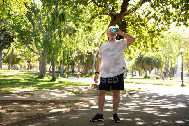 Vista frontal de un anciano caucásico maduro haciendo ejercicio en un parque en un día soleado, bebiendo agua, preparándose para hacer ejercicio - foto de stock