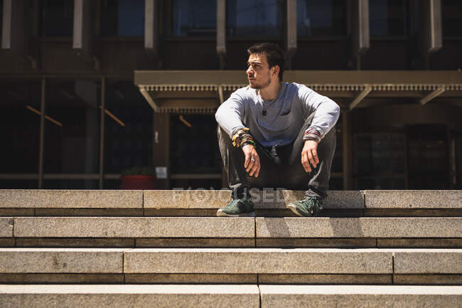 Vista frontale di un uomo caucasico che pratica il parkour vicino all'edificio in una città in una giornata di sole, si riposa prendendo una pausa, seduto sulle scale. — Foto stock
