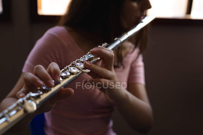 Vista lateral de cerca de una adolescente caucásica sentada frente a una ventana tocando una flauta, enfoque selectivo - foto de stock