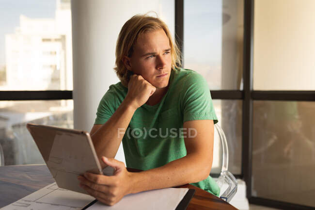 Hombre caucásico sentado junto a una mesa, usando una tableta digital y mirando hacia otro lado. Distanciamiento social y autoaislamiento en cuarentena. - foto de stock