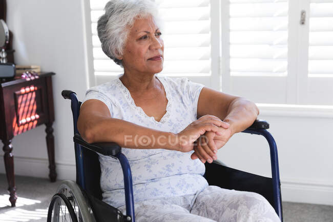 Una anciana afroamericana jubilada en casa, sentada en una silla de ruedas con pijama frente a una ventana en un día soleado mirando hacia otro lado en el pensamiento, auto aislándose durante la pandemia de coronavirus covid19 - foto de stock