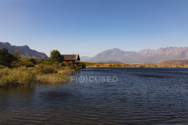 Vista de tirar o fôlego de uma cabana de madeira solitária na margem de um lago, perto das montanhas, em um dia ensolarado — Fotografia de Stock