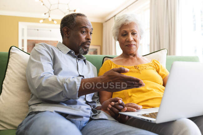 Close-up de um casal afroamericano aposentado feliz sênior em casa sentado em um sofá em sua sala de estar, conversando e usando um computador portátil juntos e sorrindo, casal isolando durante coronavírus covid19 pandemia — Fotografia de Stock