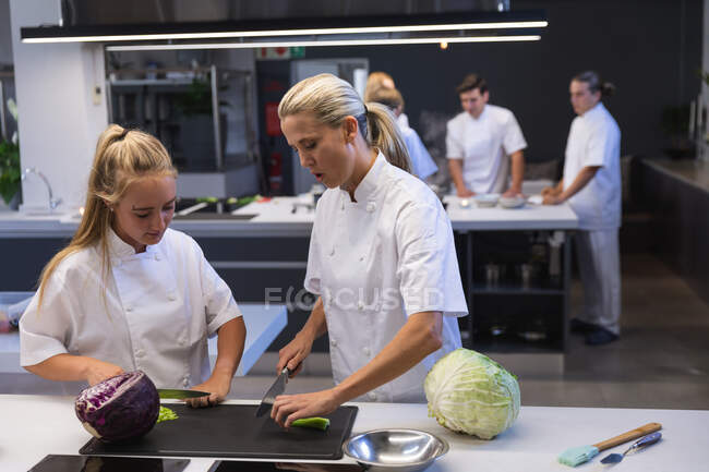 Deux femmes chefs caucasiennes coupant des légumes, parlant entre elles, avec d'autres chefs cuisinant en arrière-plan. Cours de cuisine dans une cuisine de restaurant. — Photo de stock