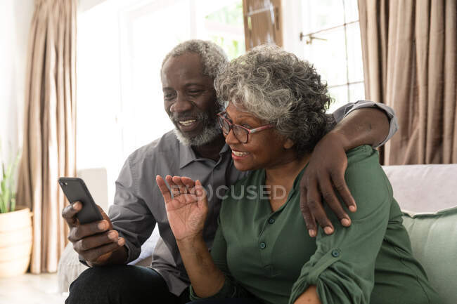 Um casal afro-americano sênior passando um tempo em casa juntos, o homem segurando um smartphone e tirando uma selfie, fazendo uma chamada de vídeo para amigos ou parentes, acenando — Fotografia de Stock