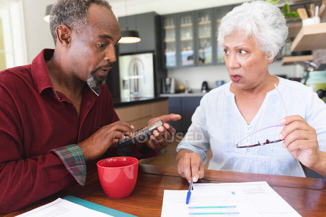 A senior retired African American couple sitting at a table in their dining room drinking coffee, looking at paperwork and discussing their finances, at home together isolating during coronavirus covid19 pandemic — Stock Photo