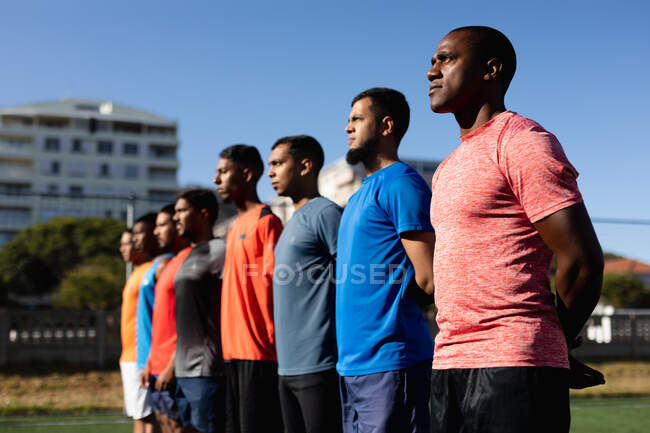 Groupe multi ethnique de cinq hommes un joueur de football latéral portant des vêtements de sport de formation sur un terrain de sport au soleil, debout dans une rangée avant un match. — Photo de stock