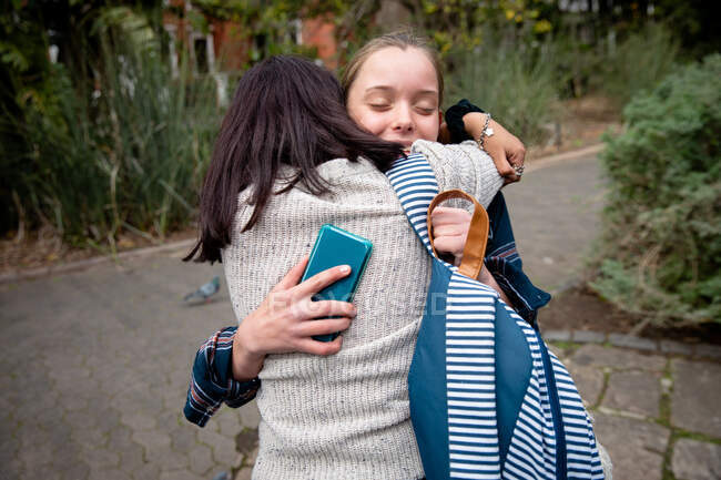 Передній вид на кавказьку і змішану расу дівчат, які проводять час разом в сонячний день, стоячи на тротуарі, обіймаючи, дівчинка тримає смартфон в руці. — стокове фото