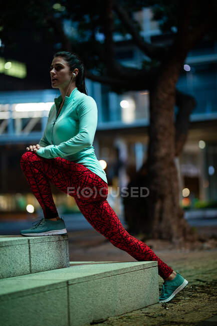 Side view of a fit Caucasian woman with long dark hair wearing sportswear exercising outdoors in the city in the evening, warming up, stretching with earphones on with urban buildings in the background. — Stock Photo