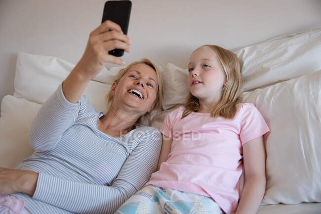 Vista frontale di una donna caucasica che si gode il tempo in famiglia con la figlia a casa insieme, sdraiata sul letto nella loro camera da letto, sorridente e scattare selfie con il suo smartphone — Foto stock