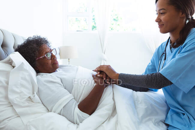 Старша змішана жінка проводить час вдома, лежачи в ліжку, відвідуючи змішану расу медсестру-жінку — стокове фото