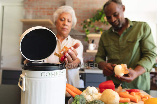 Счастливая пожилая афроамериканская пара дома, готовит еду, режет овощи, кладет овощные отходы в компостный контейнер на своей кухне, дома вместе изолирует во время пандемии коронавируса — стоковое фото