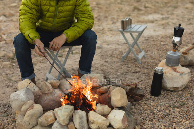 Vista frontal sección media del hombre pasar un buen rato en un viaje a las montañas, sentado junto a una fogata, viendo el fuego - foto de stock