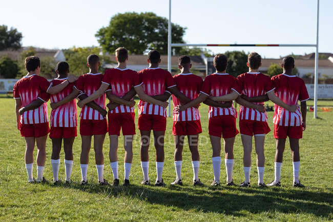 Vista trasera de un grupo de jugadores de rugby masculinos multiétnicos adolescentes que usan tira de equipo roja y blanca, de pie en un campo de juego, abrazándose. - foto de stock