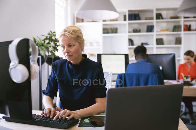 A Caucasian businesswoman working in a modern office, sitting at a desk and using a computer, with her business colleagues working in the background — Stock Photo
