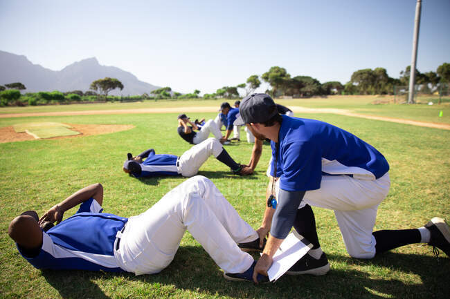 Side view of a Caucasian male baseball coach holding feet of a mixed race baseball player, instructing a multi-ethnic group of male baseball players, training before a game, working out doing sit ups at a playing field on a sunny day — Stock Photo