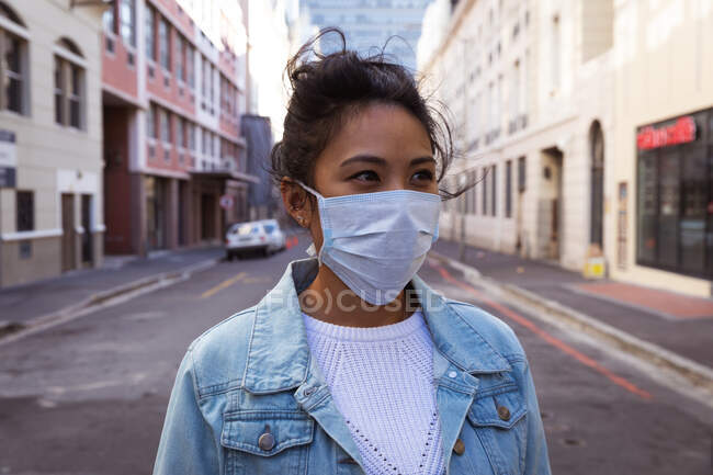 Front view of a mixed race woman with long dark hair out and about in the city streets during the day, wearing a face mask against air pollution and coronavirus, standing in a city street with buildings in the background. — Stock Photo