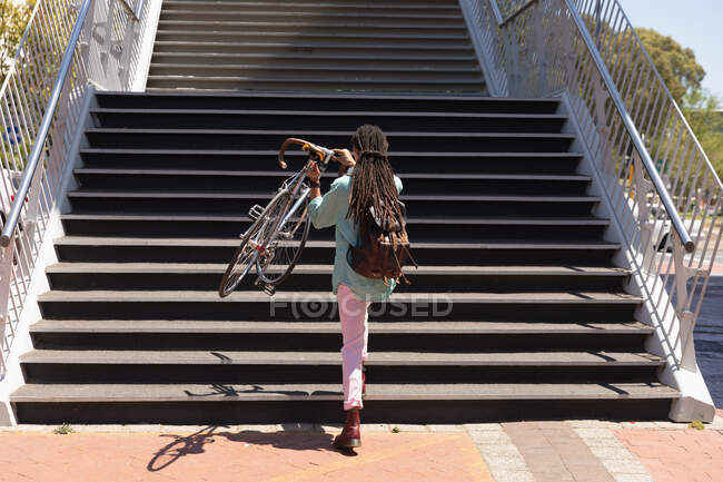 Rear view of a mixed race man with long dreadlocks out and about in the city on a sunny day, going upstairs holding his bicycle — Stock Photo
