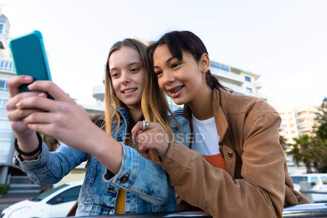 Vista frontal de una caucásica y una raza mixta chicas disfrutando del tiempo juntos en un día soleado, de pie y apoyado en la barandilla, chica tomando selfie de sí misma y su amigo. - foto de stock