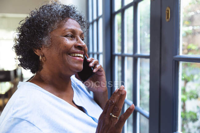 Femme de race mixte âgée profitant de son temps à la maison, de la distance sociale et de l'isolement personnel en quarantaine, regardant par une fenêtre, parlant sur un smartphone — Photo de stock