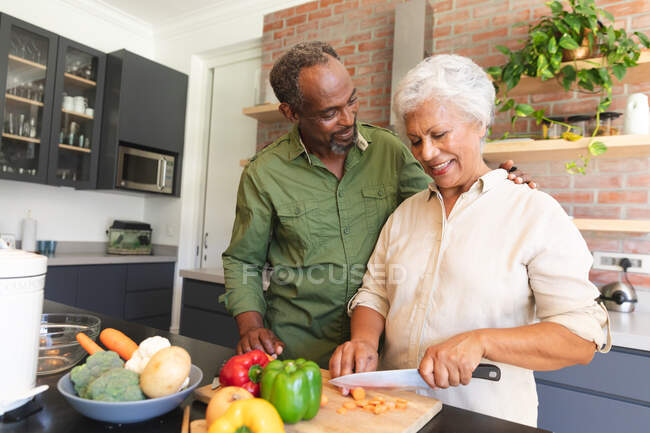 Felice anziano pensionato coppia afroamericana a casa, preparando il cibo e sorridendo nella loro cucina, la donna tagliare le verdure, l'uomo con il braccio intorno alla spalla, a casa insieme isolando durante il coronavirus covid19 pandemia — Foto stock