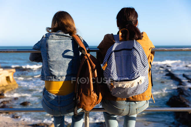 Vista posteriore di un caucasico e di una razza mista ragazze godendo il tempo insieme in una giornata di sole, seduti sulla recinzione in una passeggiata sul mare, indossando zaini. — Foto stock