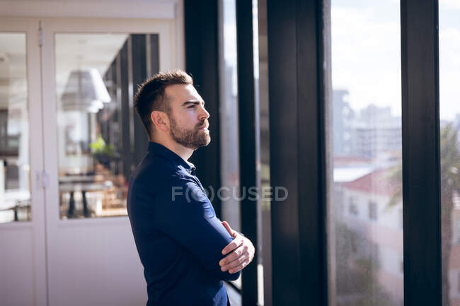 Ein kaukasischer Geschäftsmann, der in einem modernen Büro arbeitet, durch ein Fenster blickt, die Arme verschränkt und an einen sonnigen Tag denkt — Stockfoto