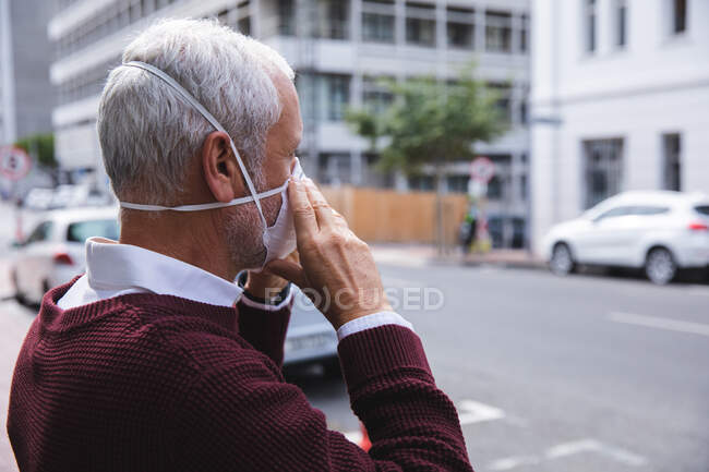 Älterer kaukasischer Mann, der tagsüber auf den Straßen der Stadt unterwegs ist und eine Gesichtsmaske gegen Coronavirus aufsetzt, covid 19 — Stockfoto