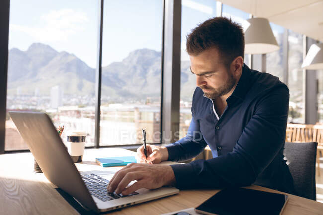 Um homem de negócios caucasiano trabalhando em um escritório moderno, sentado em uma mesa por uma janela, usando um computador portátil e escrevendo com uma caneta, tomando notas — Fotografia de Stock