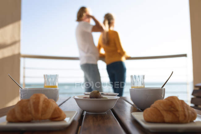 Pequeno-almoço deitado em uma mesa com casal caucasiano em pé em uma varanda, abraçando ao fundo. Distanciamento social e auto-isolamento em quarentena. — Fotografia de Stock