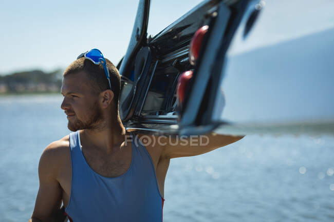 Передній вид упевненого кавказького веслувальника в блакитній футболці з човном на плечі, що йде через річку в сонячний день. — стокове фото