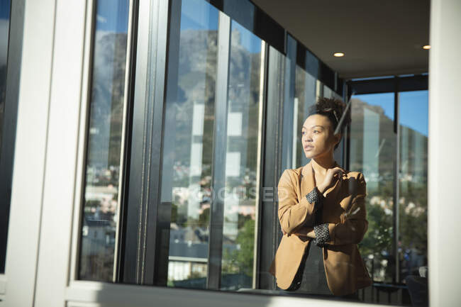 Eine Geschäftsfrau mit gemischter Rasse, die in einem modernen Büro arbeitet, durch ein Fenster blickt und an einem sonnigen Tag mit verschränkten Armen denkt — Stockfoto