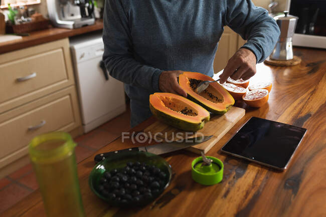 Vista lateral sección media del hombre relajándose en casa, de pie en el mostrador en su cocina preparando cuidadosamente un melón partido a la mitad con una cuchara antes de comer - foto de stock