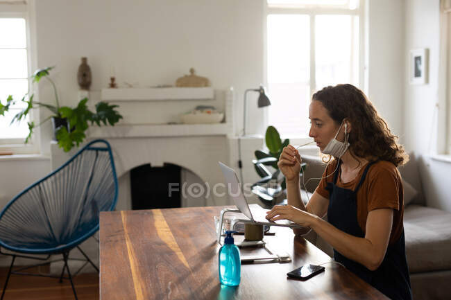 Mujer caucásica pasando tiempo en casa, usando una máscara facial contra coronavirus, covid 19, sentada junto a su escritorio y trabajando, usando su portátil y auriculares. Distanciamiento social y autoaislamiento en cuarentena. - foto de stock