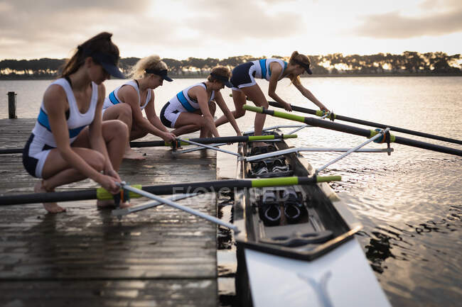 Side view of a rowing team of four Caucasian women on a jetty preparing the oars on a boat on the river for rowing training — Stock Photo