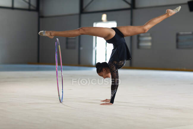 Vue latérale de gymnaste mixte adolescente performant au gymnase, faisant de l'exercice avec un cerceau, se tenant à la main et se divisant, portant un justaucorps noir — Photo de stock