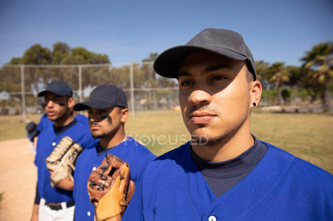 Vista frontal de un grupo multiétnico de jugadores de béisbol, preparándose antes de un partido, de pie en fila, escuchando un himno nacional - foto de stock