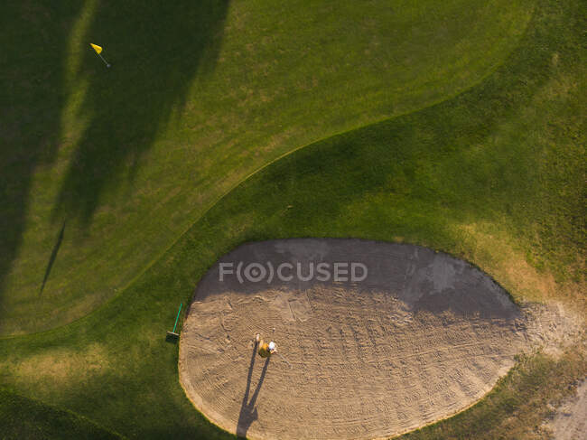 Drone shot of a man playing golf at a golf course on a sunny day, standing in a bunker aiming for the hole — Stock Photo