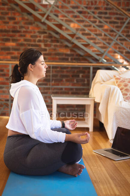 Vlogger femenina caucásica en casa en su sala de estar, practicando yoga y usando su computadora portátil. Distanciamiento social y autoaislamiento en cuarentena. - foto de stock