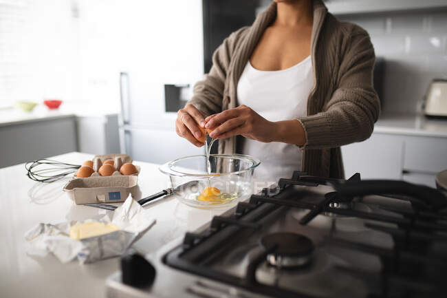 Вид спереди на среднюю часть женщины дома, стоящей на кухне у стола, готовящей завтрак, разбивающей яйцо в миску — стоковое фото
