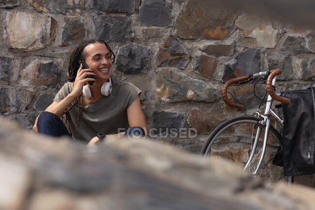 Вид спереду змішаного гоночного чоловіка з довгими сновидіннями і близько в місті в сонячний день, сидячи біля стіни на вулиці і посміхаючись, використовуючи смартфон, з велосипедом, що нахиляється навпроти стіни поруч з ним . — стокове фото