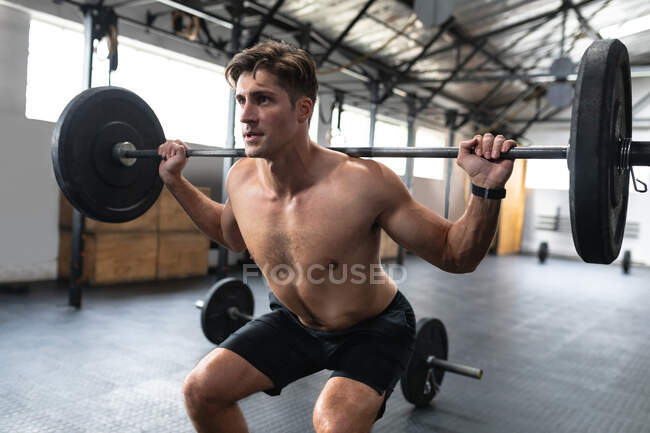 Front view of a shirtless athletic Caucasian man cross training at a gym, weight training with barbells, standing with bent legs and the weights on his shoulders, focusing before the final lift — Stock Photo