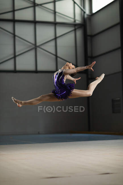 Бічний вид підлітка-кавказької жіночої гімнастки виступає в спортзалі, стрибаючи і роблячи розкол, одягаючи фіолетовий летар. Гімнасти наполегливо тренуються для змагань. — стокове фото